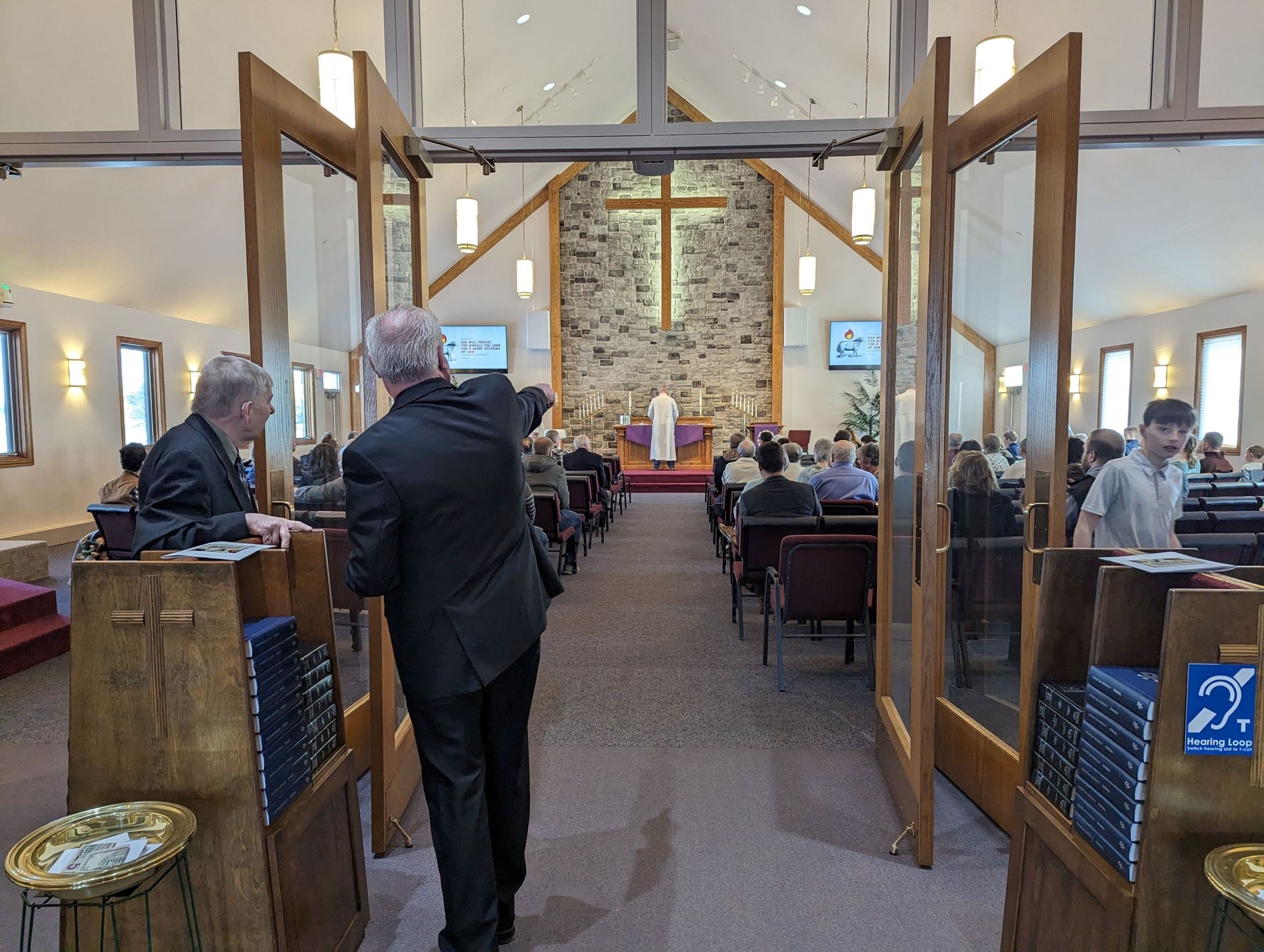 Interior of a church with an open entrance, people seated, and a person at the altar.