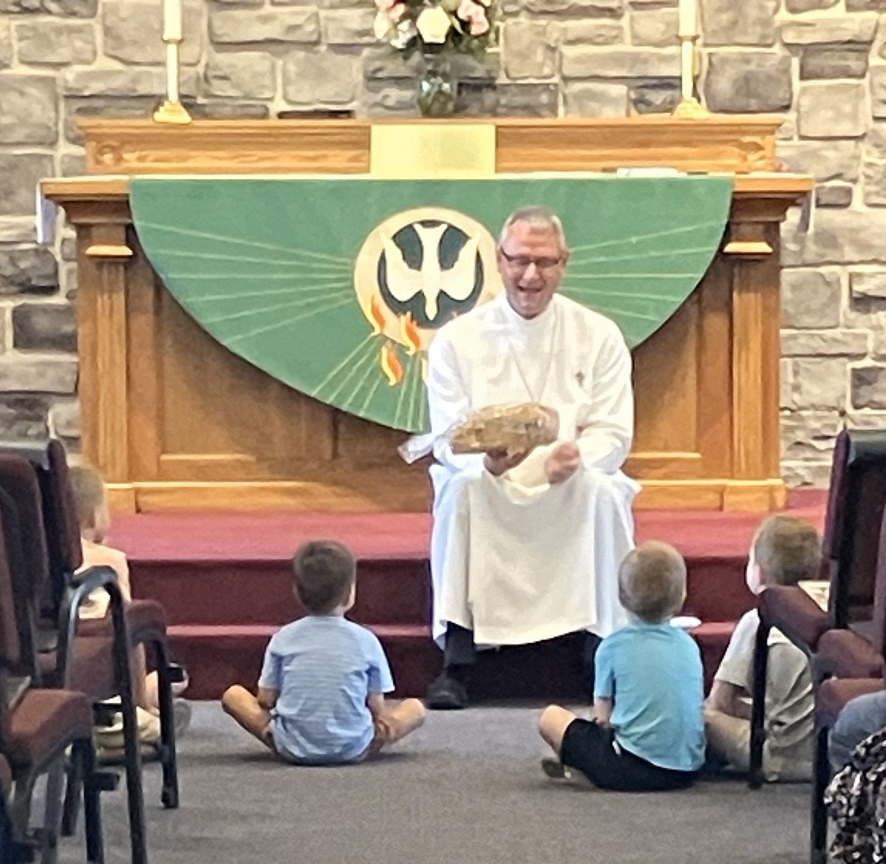 Clergyman in white robe sitting on altar steps with children, holding a loaf of bread.