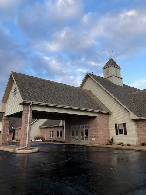 Exterior of a church with a steeple and cross, under a cloudy blue sky.